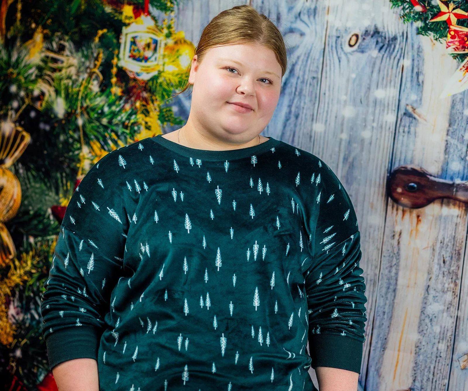 A young person with light skin and blonde hair, wearing a dark sweatshirt with a small tree pattern, standing in front of a decorated Christmas tree and a wooden background.