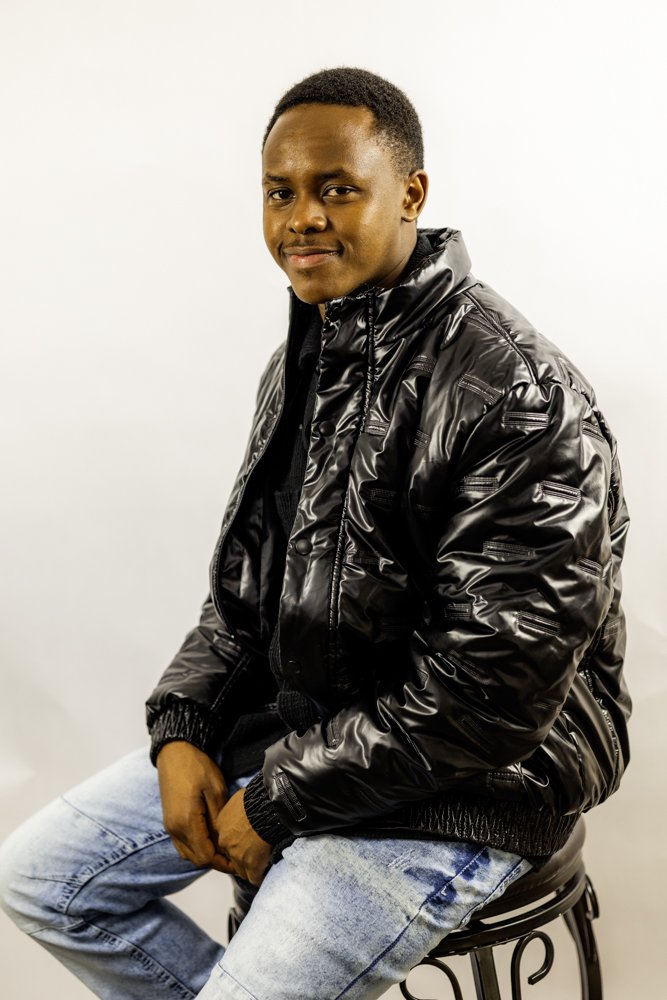 Young man sitting on a black stool, wearing a shiny black bomber jacket and light blue jeans, smiling at the camera against a plain white background.