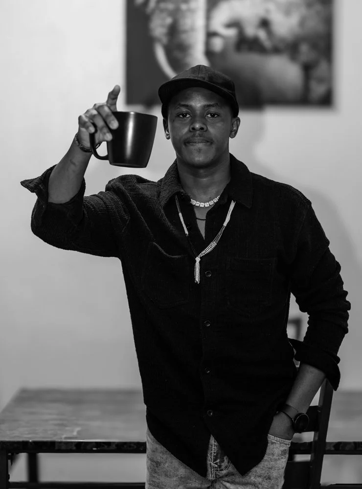 A young man wearing a black baseball cap, necklaces, and a watch, holding a mug in his right hand, standing indoors with a blurred wall art in the background.
