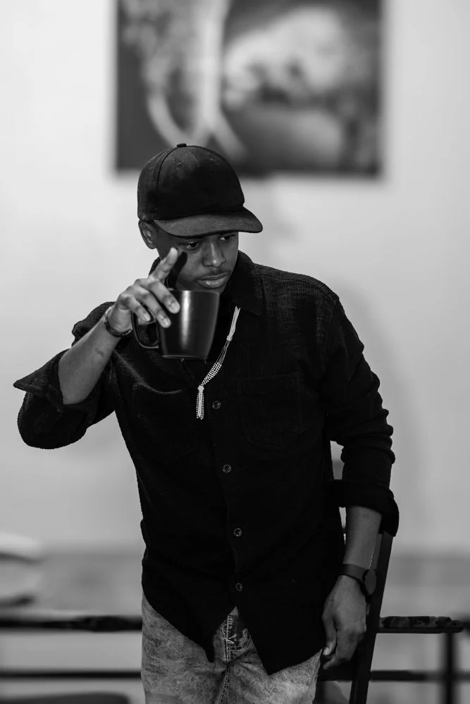 A young man in a black baseball cap and dark shirt holding a black mug, looking down thoughtfully, with a blurred globe picture on the wall behind him.