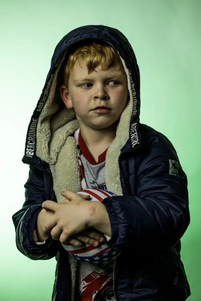 Young boy with red hair wearing a navy blue jacket with a hood, holding a soccer ball, against a green gradient background.