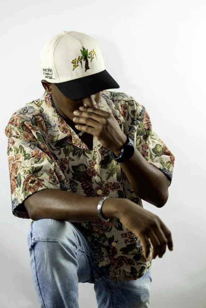 A man wearing a floral short-sleeve shirt, light blue jeans, and a baseball cap with a tree, lightning bolts, and writing on it. He is crouching, with his left elbow resting on his knee, and covering his mouth with his right hand while looking down.