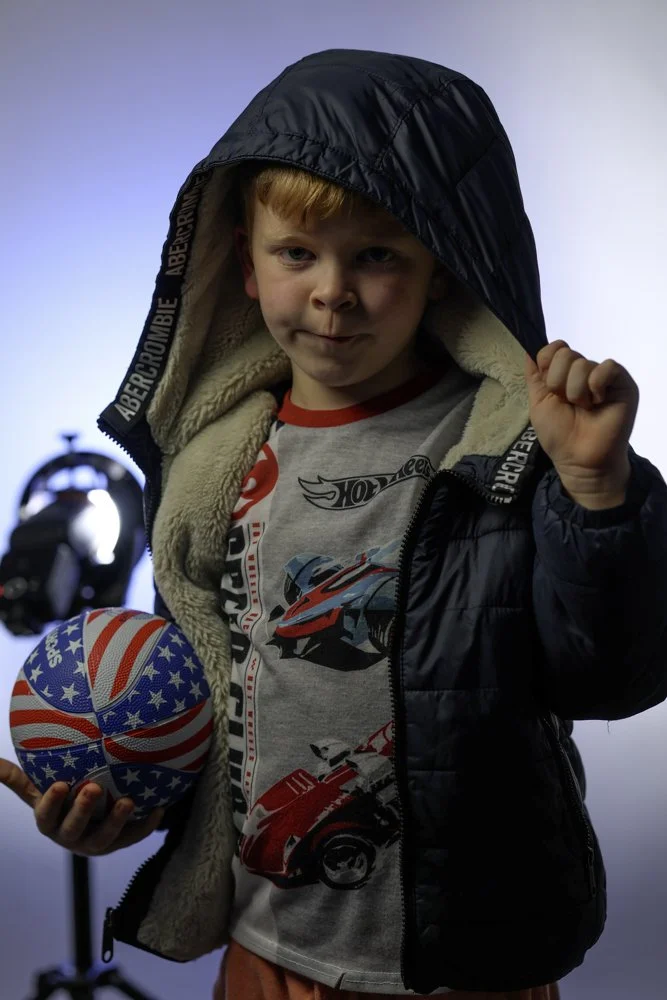 Young boy wearing a hooded jacket and a gray t-shirt with car graphics, holding a basketball decorated with stars and stripes, standing near a camera.
