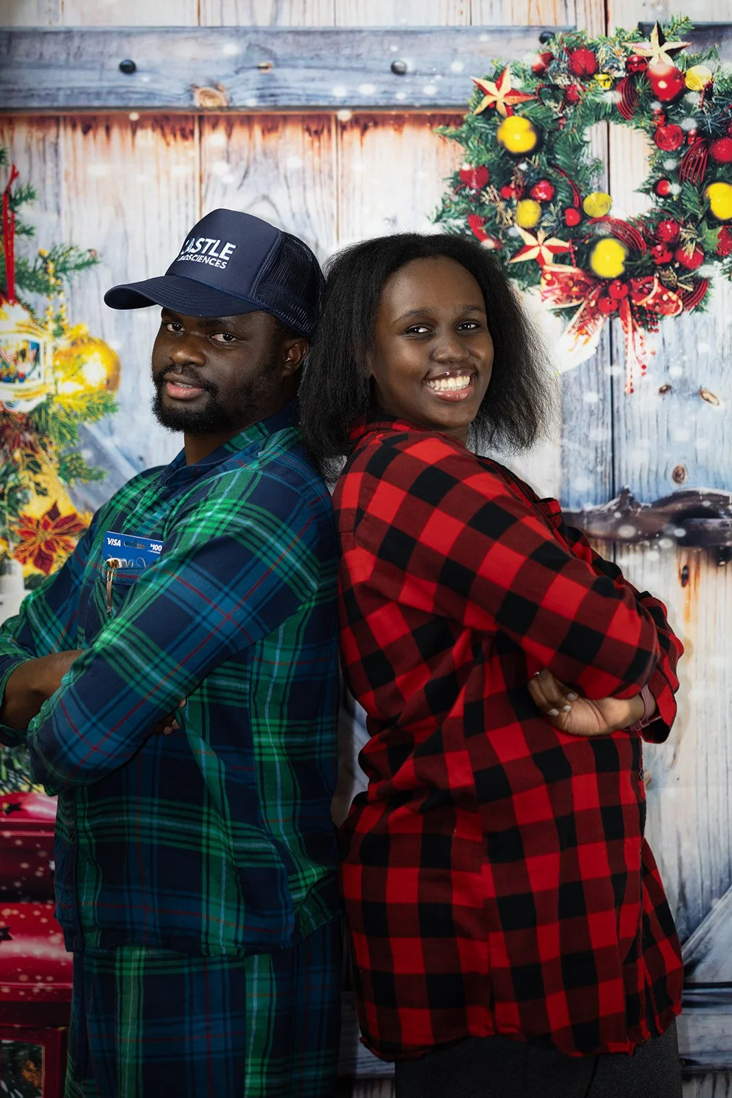 A man and woman stand back-to-back with arms crossed, smiling, in front of a Christmas holiday backdrop with wreaths and ornaments, both wearing plaid shirts.