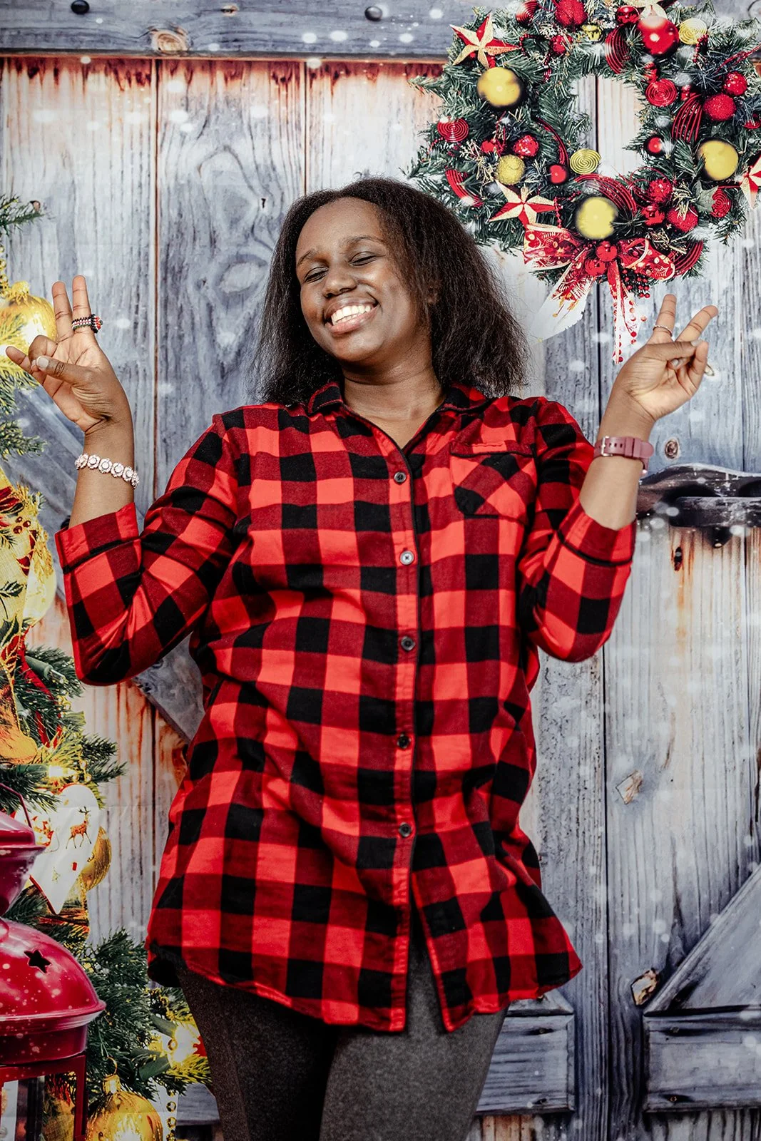 A woman smiling and posing with peace signs, wearing a red and black checkered shirt, in front of a decorated Christmas background with ornaments, a wreath, and a wooden wall.