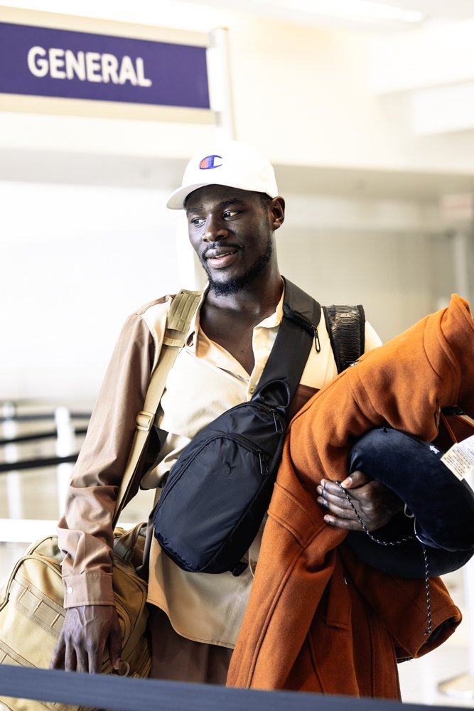 A young man at an airport baggage claim area holding a red jacket and a black cap, with a beige duffel bag and a black backpack, in front of a sign that says 'GENERAL'.