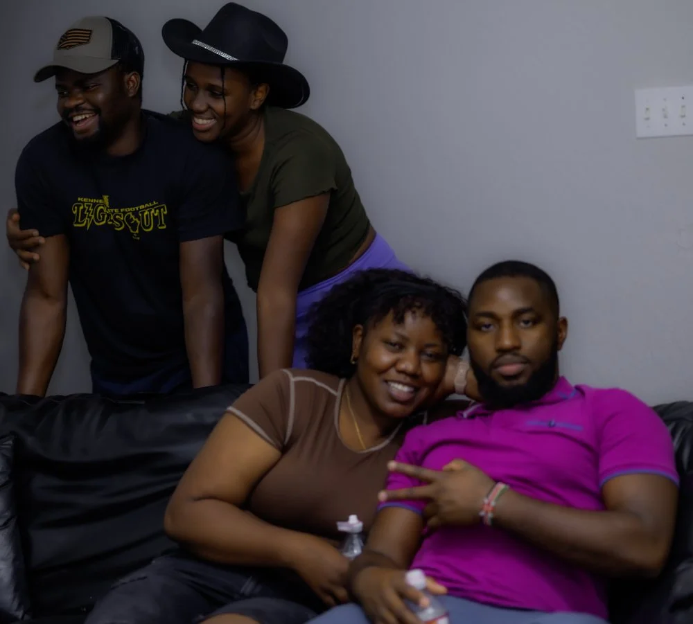 Four friends smiling and posing casually in a living room, with two sitting on a black leather couch and two standing behind them. They appear happy and relaxed.