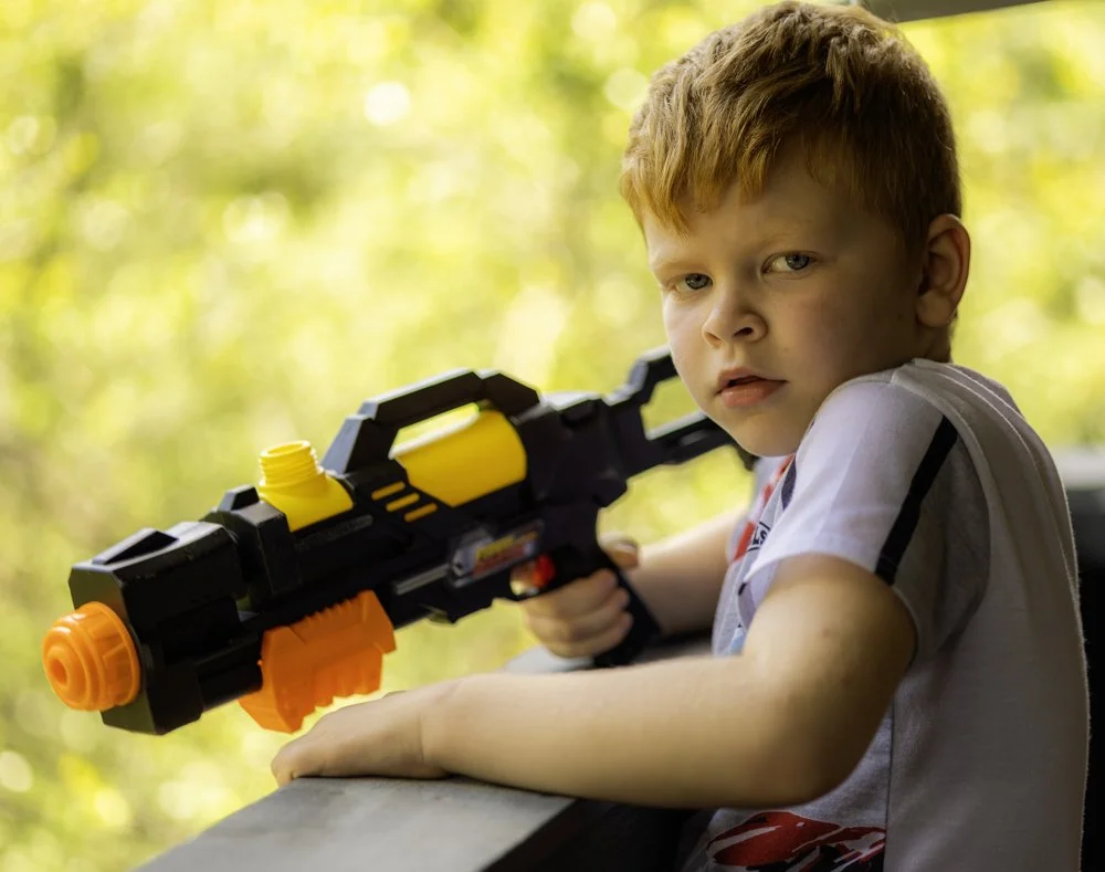 Young boy with red hair aiming a toy water gun outdoors with a blurred green background.