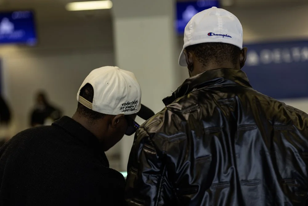 Two men at an airport, wearing hats and jackets, looking at an electronic device.