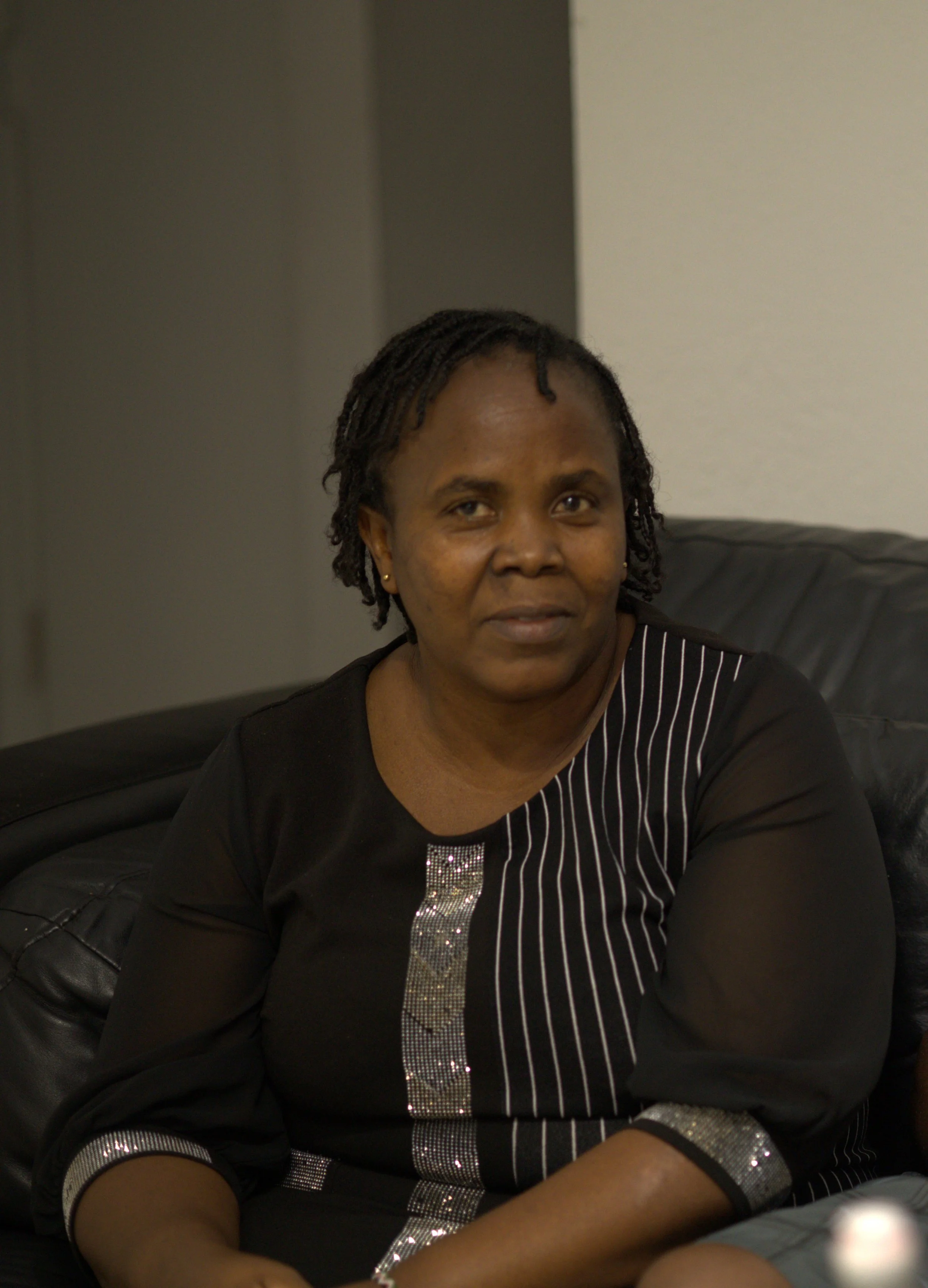 A woman with short dreadlocks sitting on a black leather couch, wearing a black top with silver accents and sheer sleeves, in a room with neutral-colored walls.