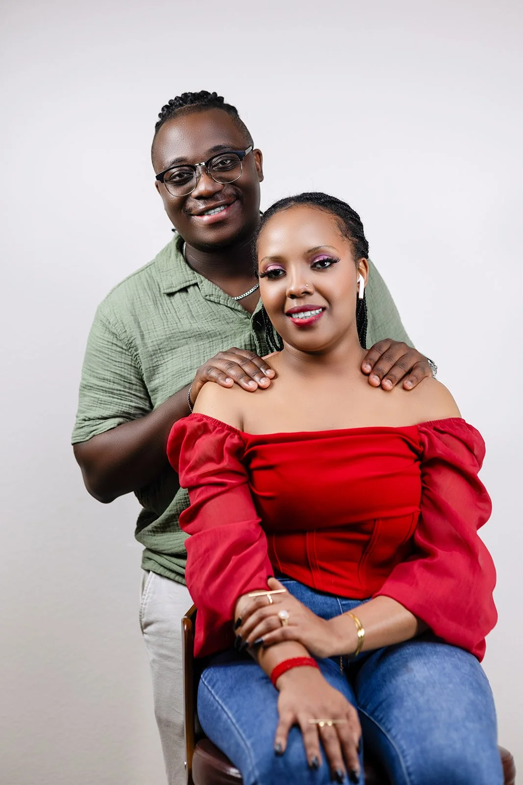 A man standing behind a woman, both smiling, with the man's hands resting on the woman's shoulders. The woman is sitting, wearing a red off-the-shoulder top, and the man is wearing glasses and a green shirt. They are in a studio setting with a plain 