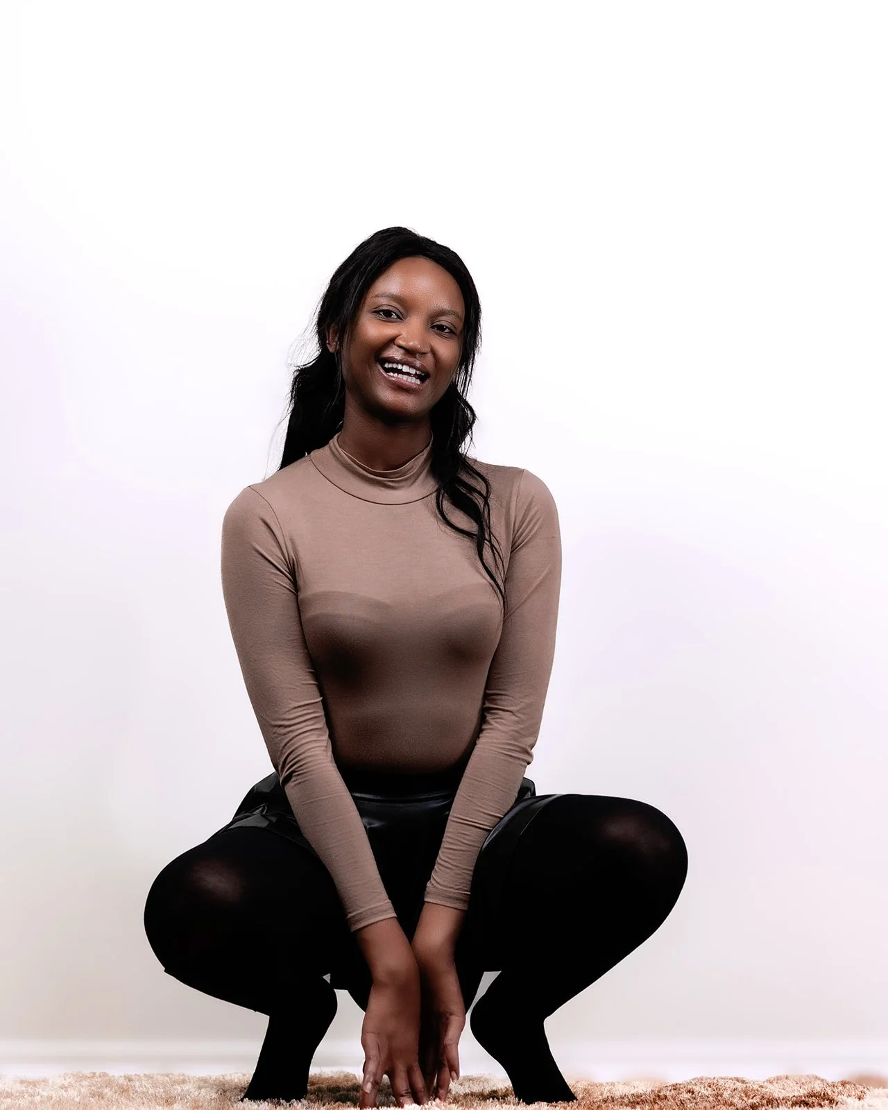 A woman with dark hair smiling and squatting on a light-colored carpet against a plain white background.