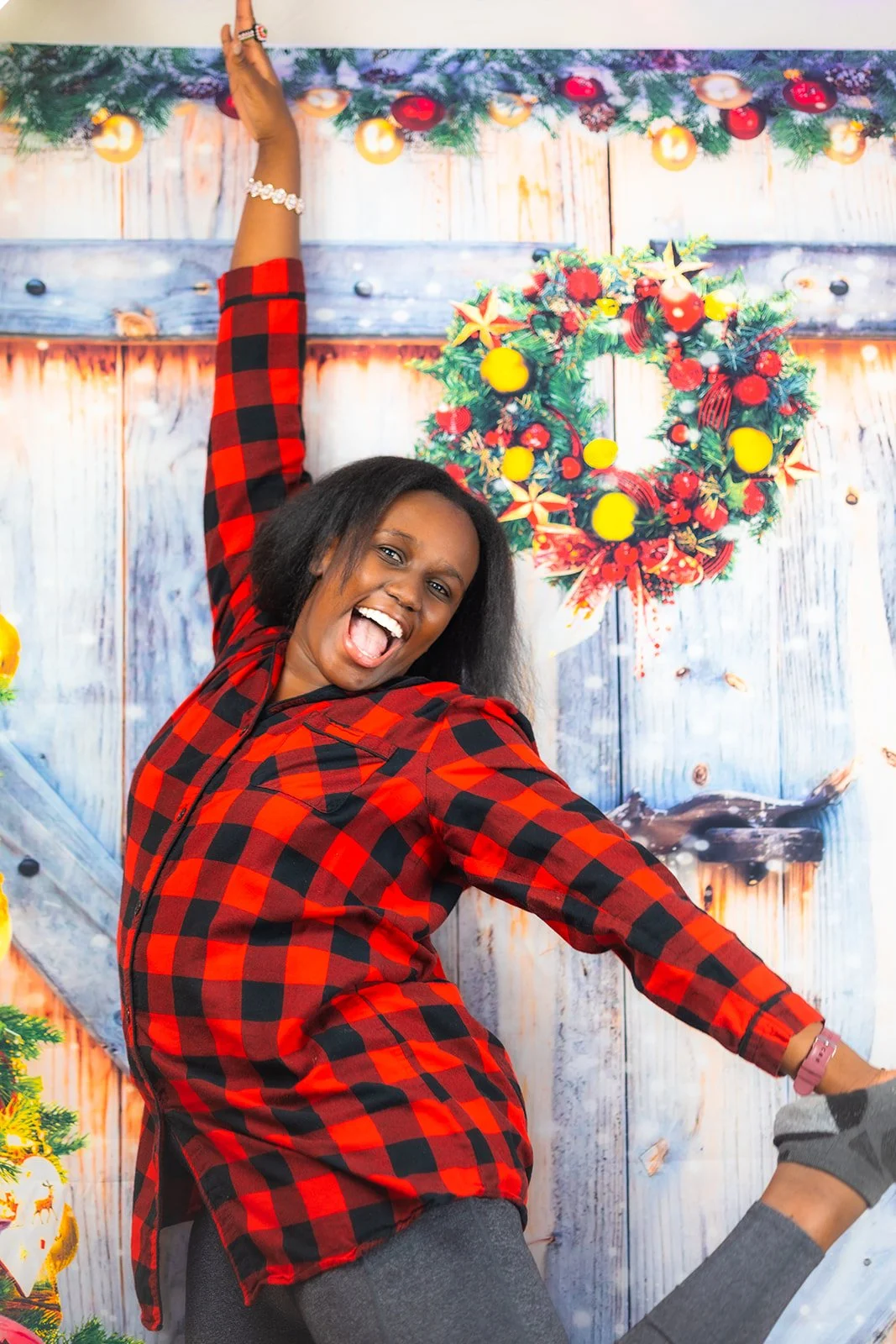 Woman in a red and black plaid shirt cheerfully posing, with festive Christmas decorations and a wreath with red and yellow ornaments in the background.