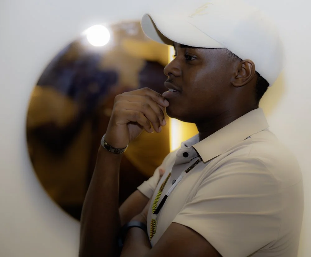 A young man in a beige cap and shirt looking into an airplane window, reflecting his face and hand in the glass.