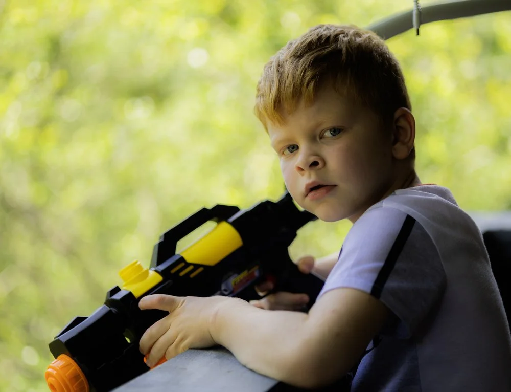 A young boy with red hair holding a toy gun outdoors with a blurred green background.