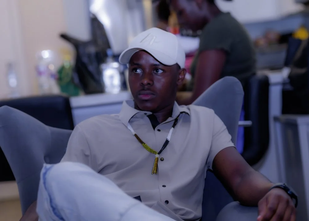 A young man sitting in a modern indoor space, wearing a white baseball cap and a beige shirt, with an attentive expression.