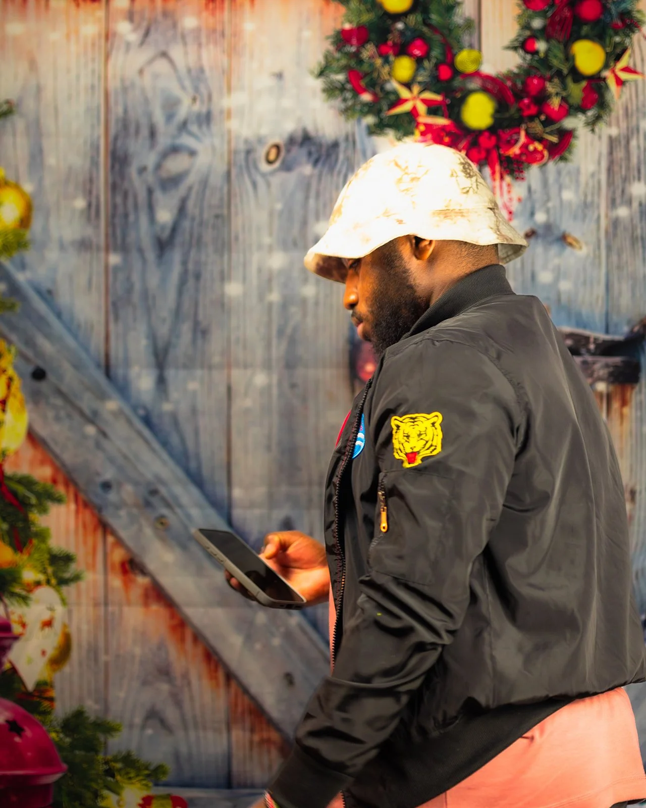 A man wearing a black jacket with patches, including a yellow tiger face, and a light-colored bucket hat, looking at his phone against a festive decorated background.