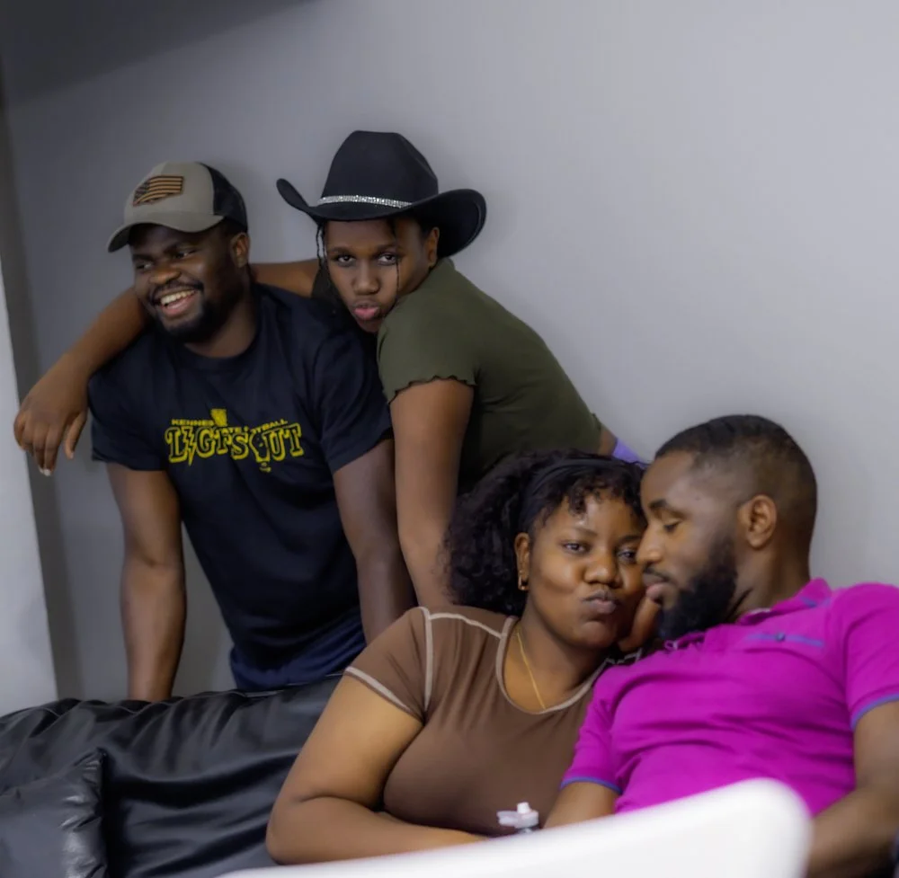 Four people in a close group; two are sitting on a couch, and two are leaning over them. One person is wearing a cowboy hat, and another is wearing a baseball cap with an American flag. The group appears happy and affectionate.