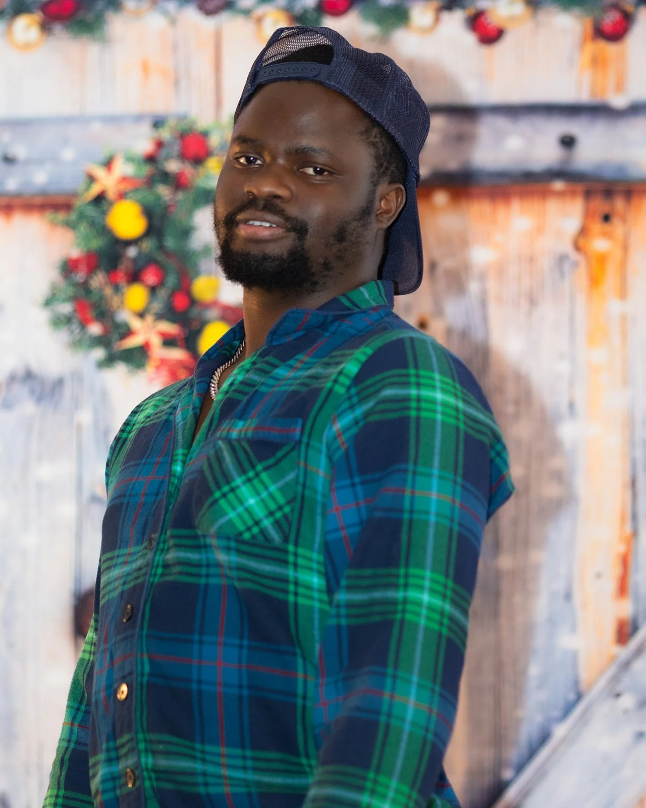 A man in a green and blue plaid shirt and a backward navy baseball cap stands indoors in front of a rustic wooden wall decorated with a holiday wreath of red, yellow, and green ornaments.