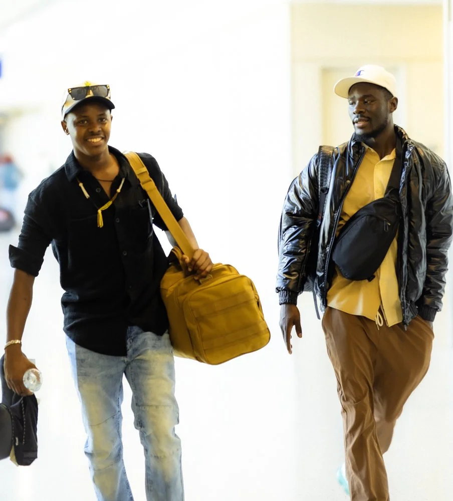Two young men with backpacks and casual clothing, walking through an airport or similar setting, smiling and engaged in conversation.