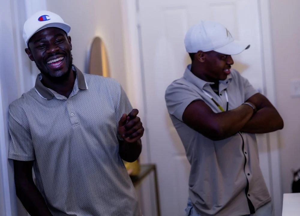 Two young men wearing white caps and casual shirts laughing and smiling indoors.