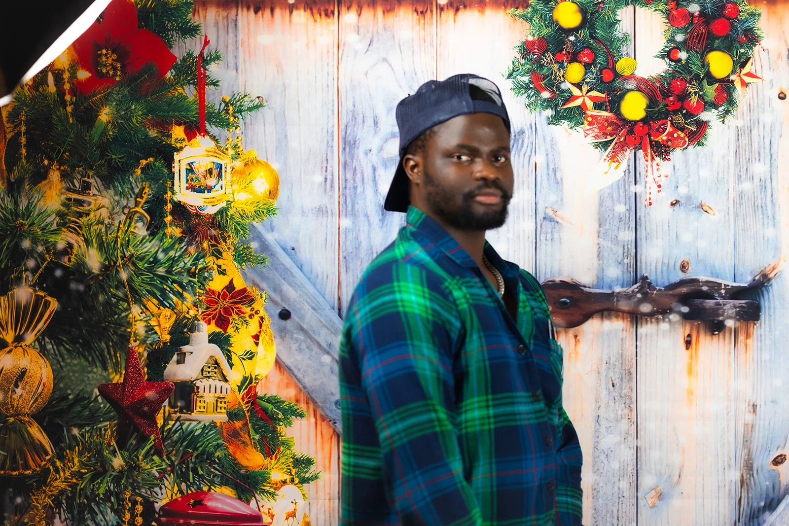 A man in a green and blue plaid shirt and a dark baseball cap standing in front of a festive Christmas backdrop. The background features a decorated Christmas tree with gold and red ornaments, a star, and other holiday decorations, along with a white