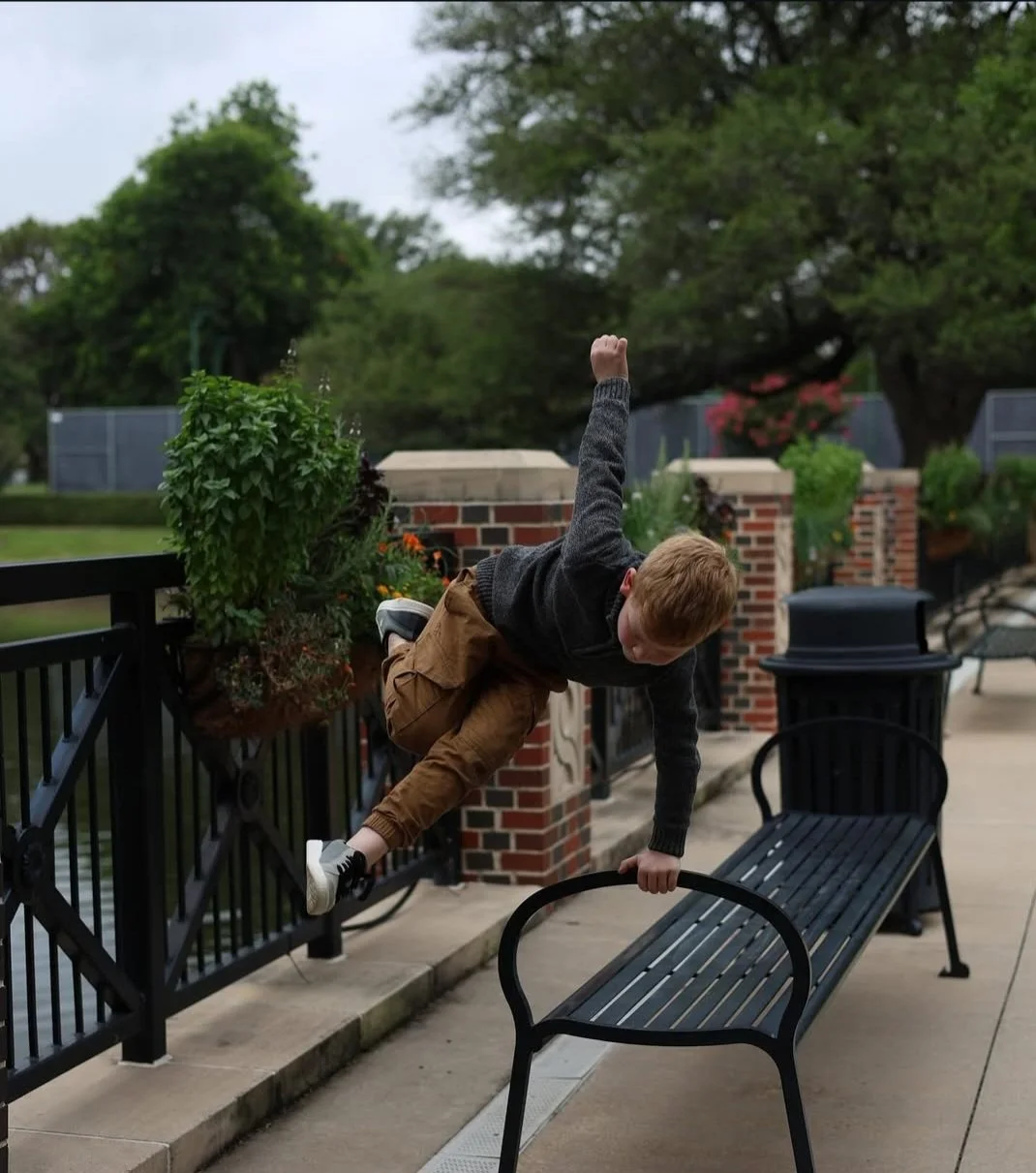 A young boy is hanging upside down from a black metal park bench, gripping the top of the backrest with one hand and the armrest with the other, as he appears to be climbing or playing football. The setting appears to be a park or outdoor area with greenery and brick walls.