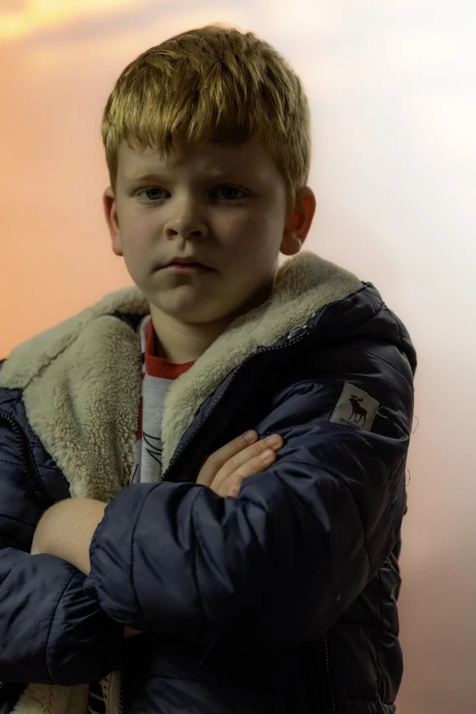 A young boy with reddish-brown hair and a serious expression, wearing a navy puffer jacket with a beige fleece collar, standing against a soft pink and orange background.