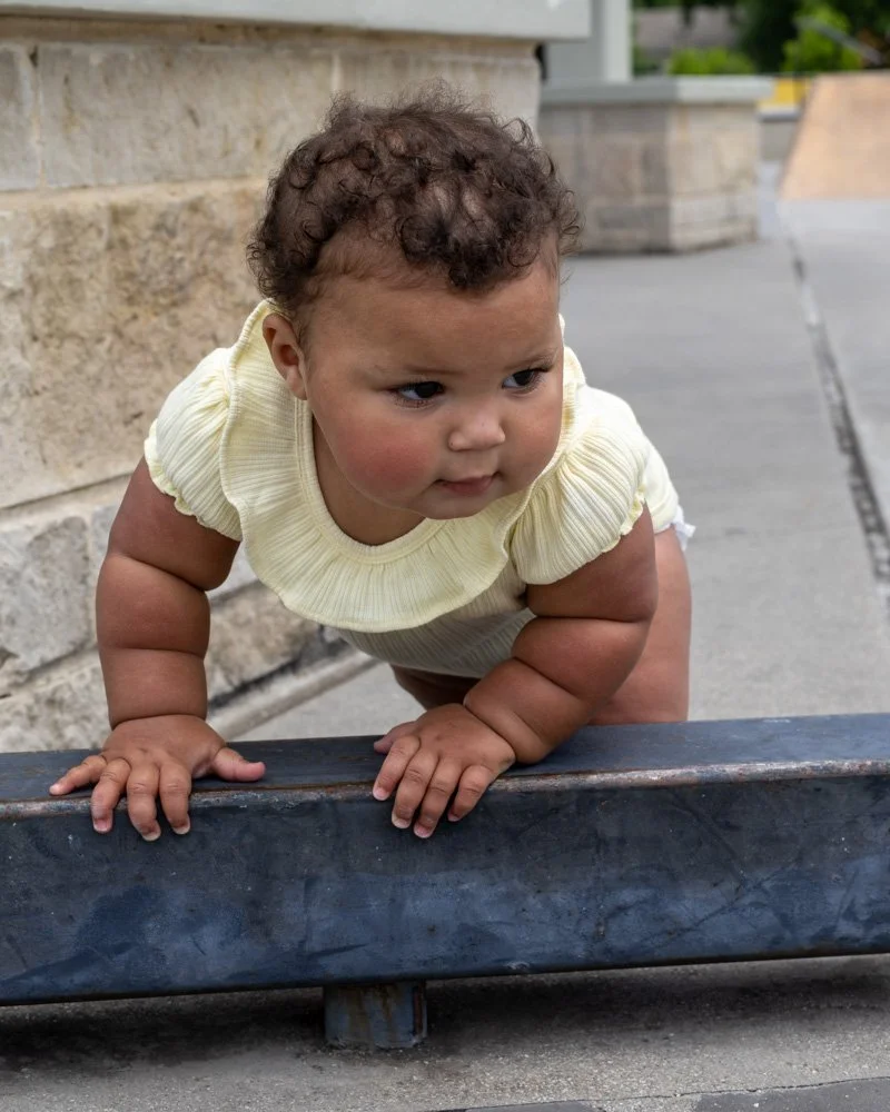 A young child with curly hair climbing over a metal barrier outdoors, wearing a yellow top with ruffled sleeves.