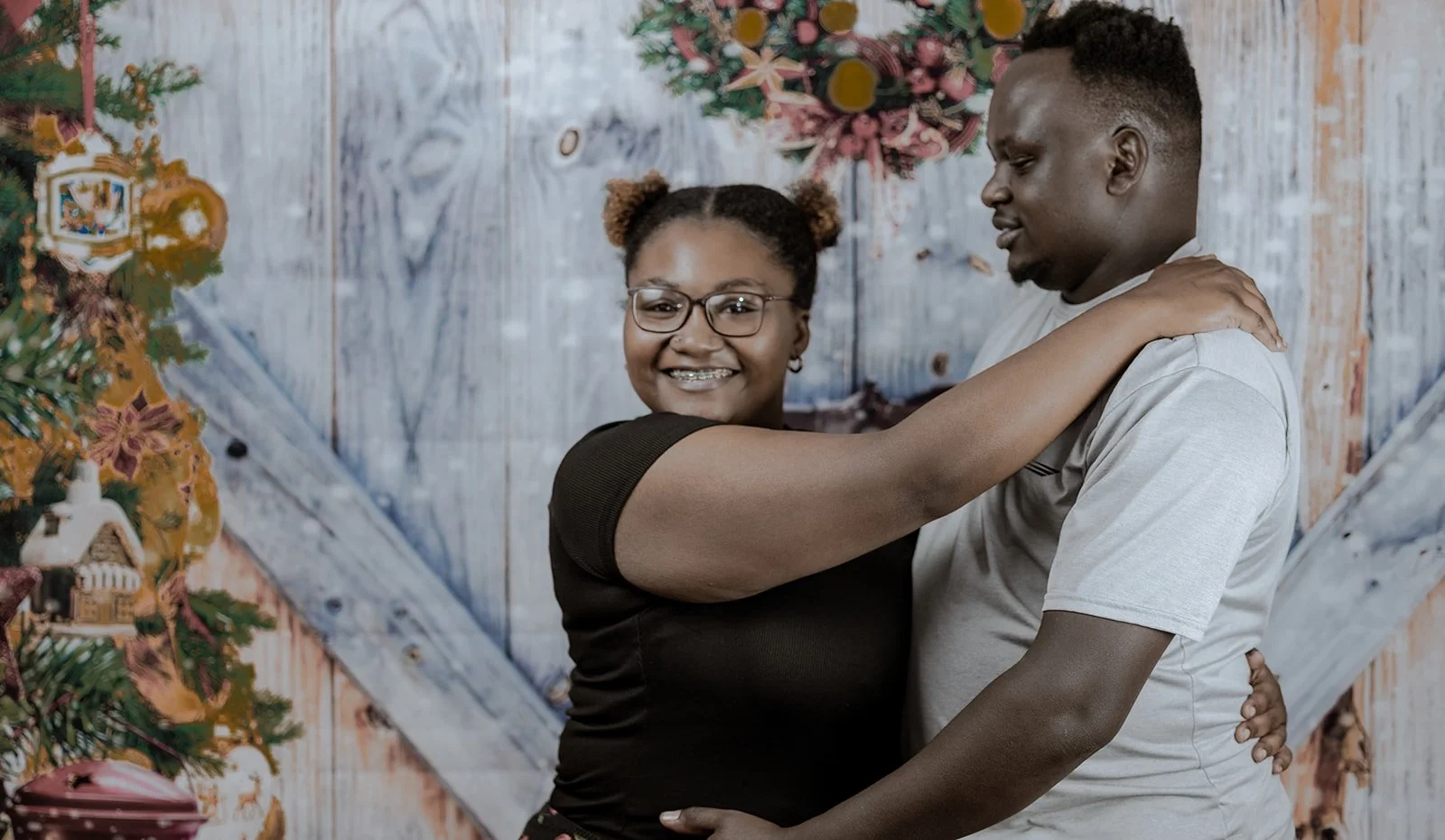 A smiling woman with glasses hugging a man in front of a Christmas decorated background.