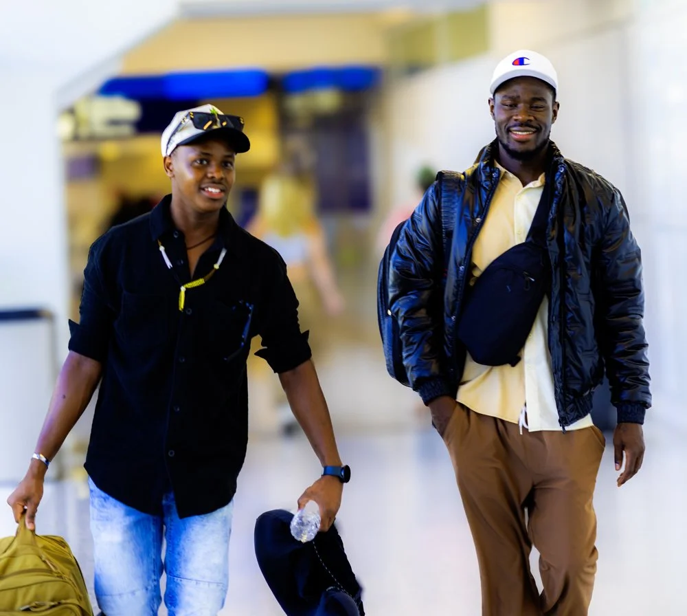Two men walking in an airport, smiling. One is holding a yellow backpack and a water bottle; the other has a black bag and backpack.
