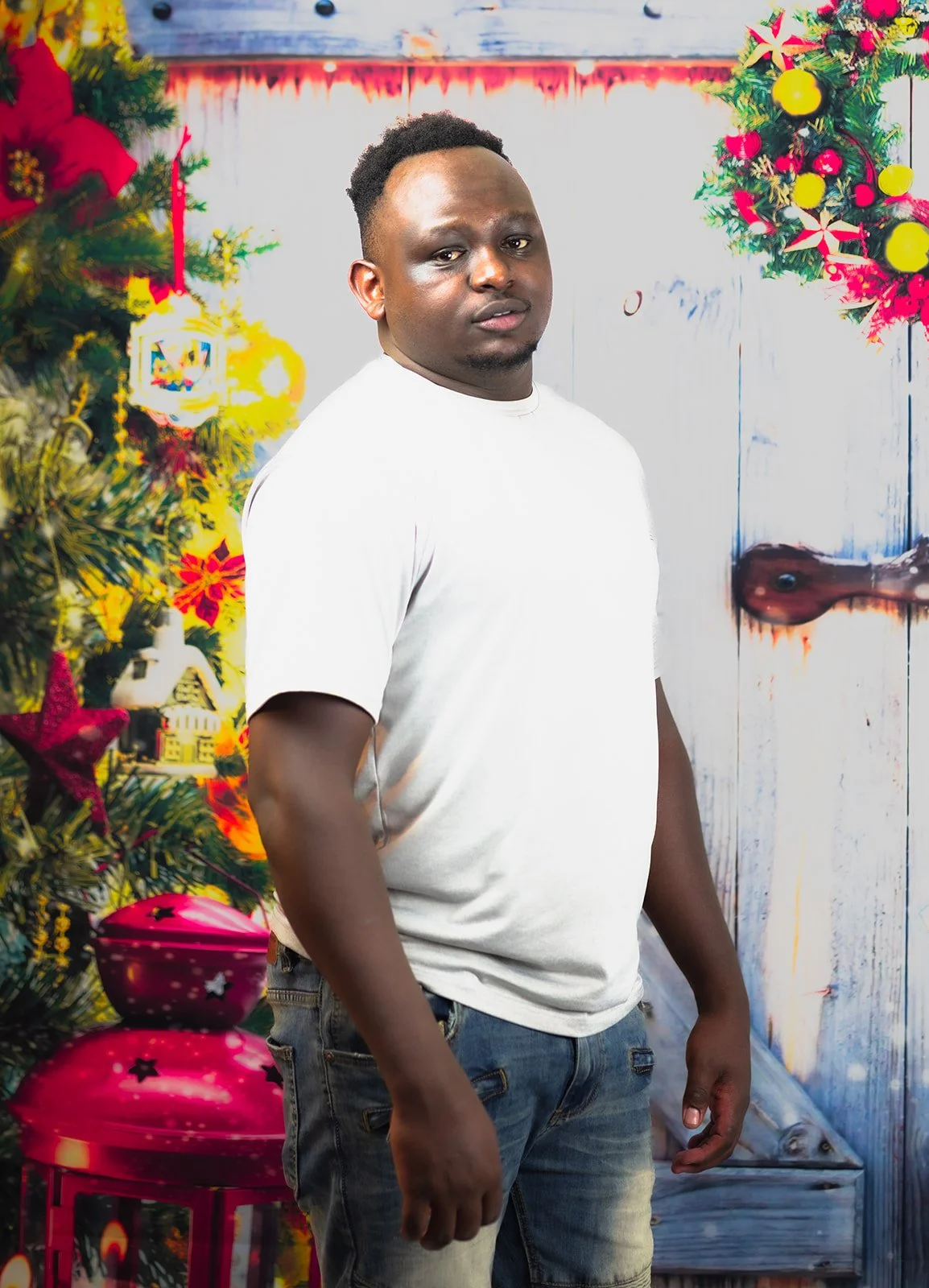 A man in a white T-shirt and jeans standing in front of a Christmas-themed background with a decorated Christmas tree, wreath, and holiday ornaments.