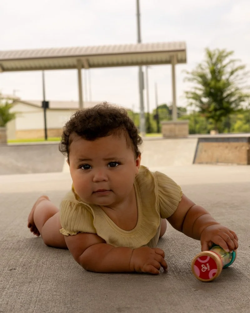A baby with curly hair lying on their stomach outdoors on a gray surface, holding a colorful toy, with a park and trees in the background.