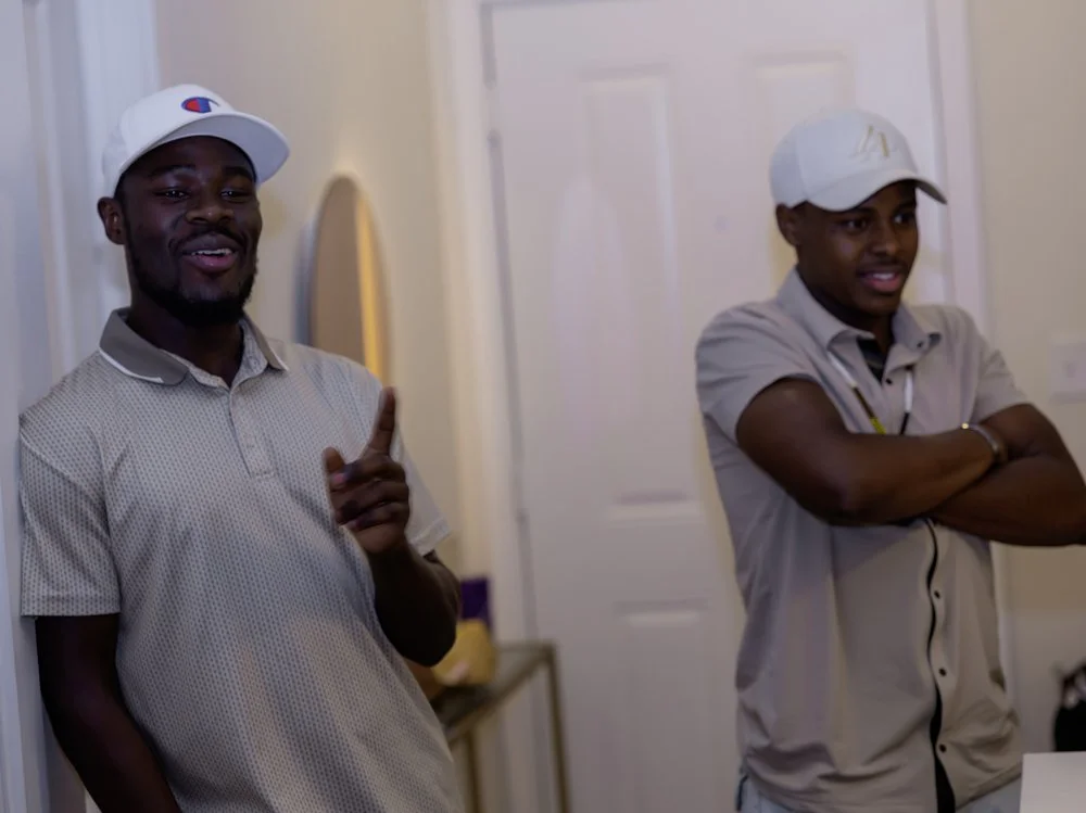 Two young men at a social gathering or party, wearing caps and casual shirts, with one smiling and gesturing while the other looks on with crossed arms.