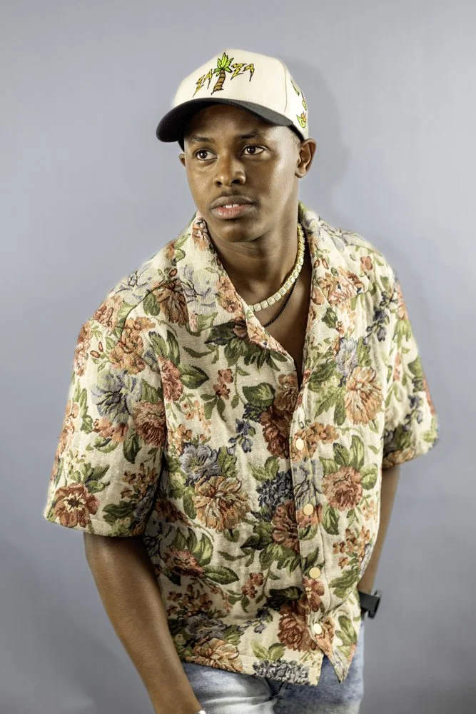 Young man in floral shirt, baseball cap, and pearl necklace against a plain gray background.