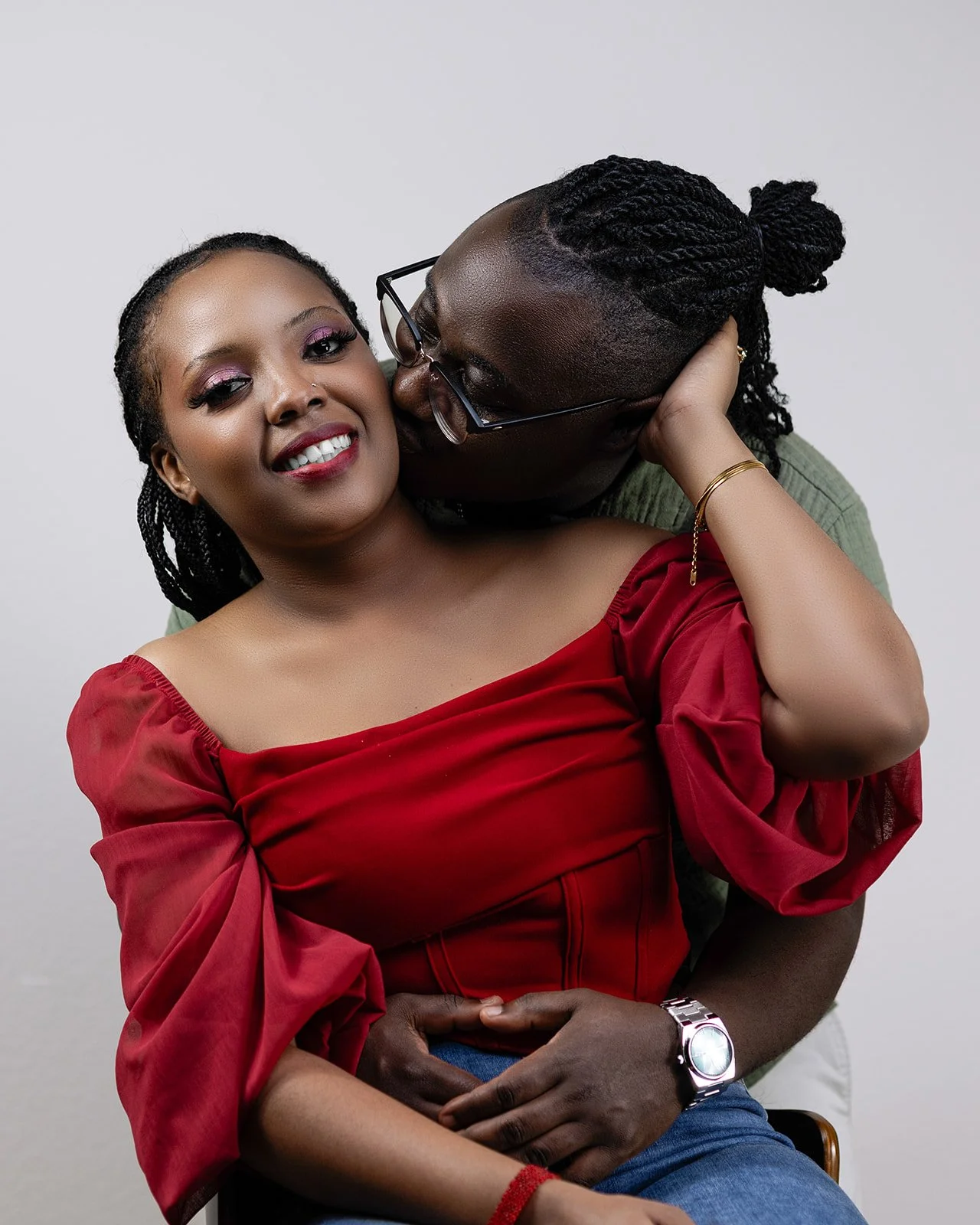 A man affectionately kisses a woman on the cheek while sitting close together against a plain background.