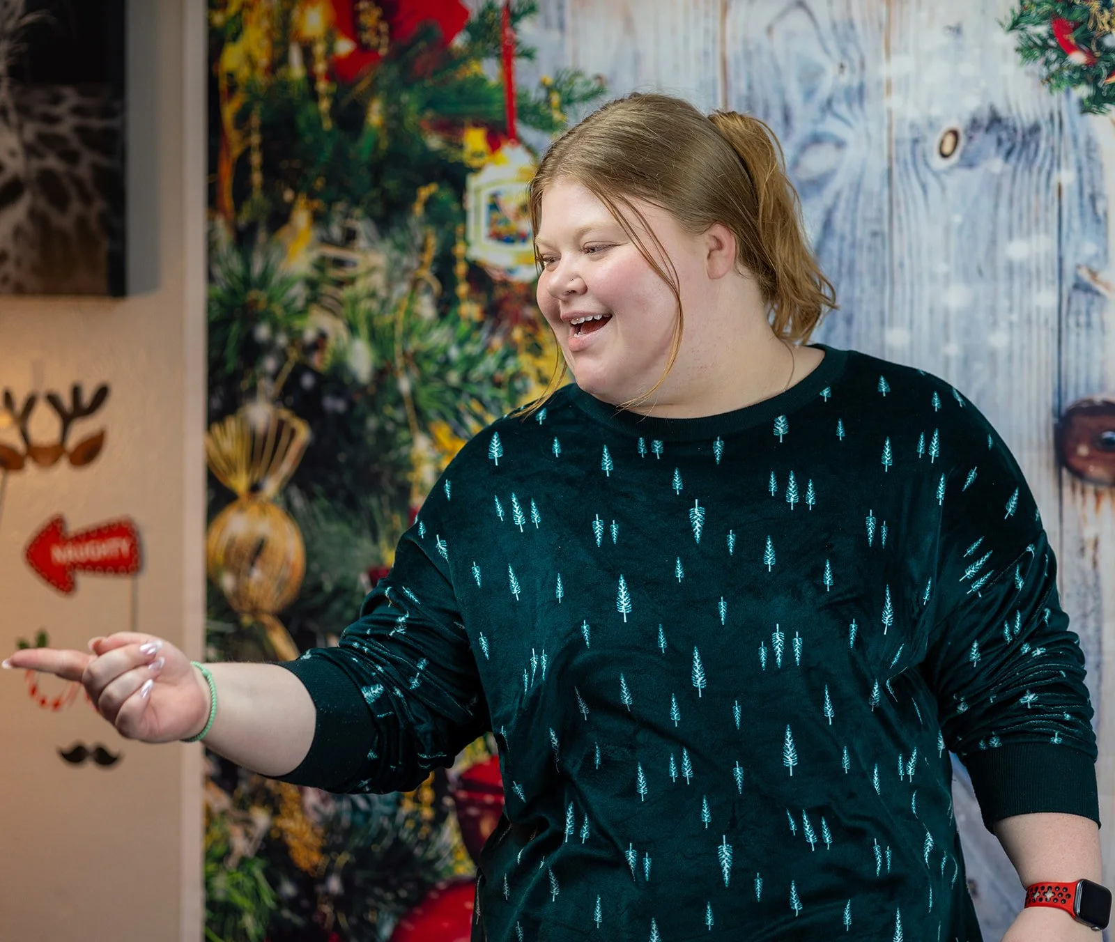 A woman with red hair smiling and pointing, wearing a black sweatshirt with blue Christmas trees, standing in front of a decorated Christmas tree and holiday-themed wall decorations.