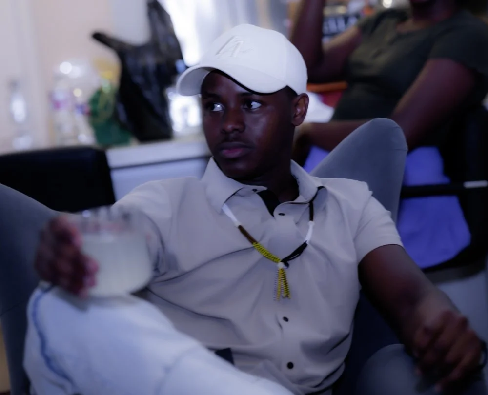 A young man wearing a white cap and shirt, sitting in a room with other people, holding a bowl of food, looking to the side.