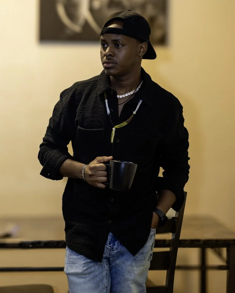 A young man wearing a black backwards cap, black shirt, and light blue jeans, holding a black mug and standing indoors with a neutral expression.