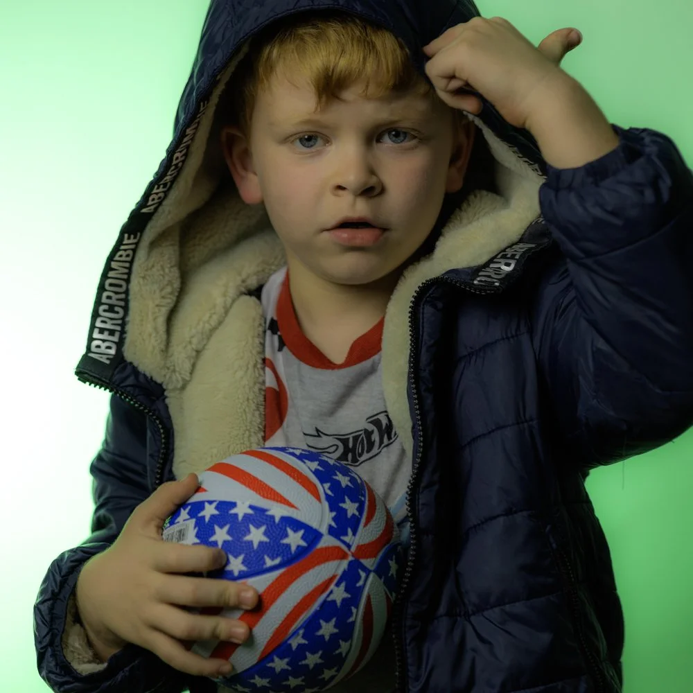 A young boy with blond hair and blue eyes, wearing a navy blue jacket with a hoodie, a white and red shirt, holding a patterned soccer ball with American flag design, against a split green background.