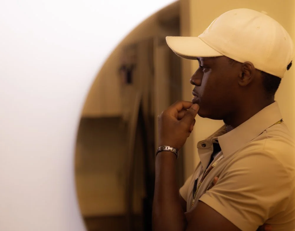 A man wearing a beige polo shirt and a white cap, standing in front of a mirror, appears to be deep in thought.