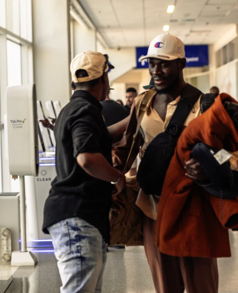 Two men talking at an airport security checkpoint, one in a black shirt and tan cap, the other in a brown jacket, white cap, and carrying a red jacket.