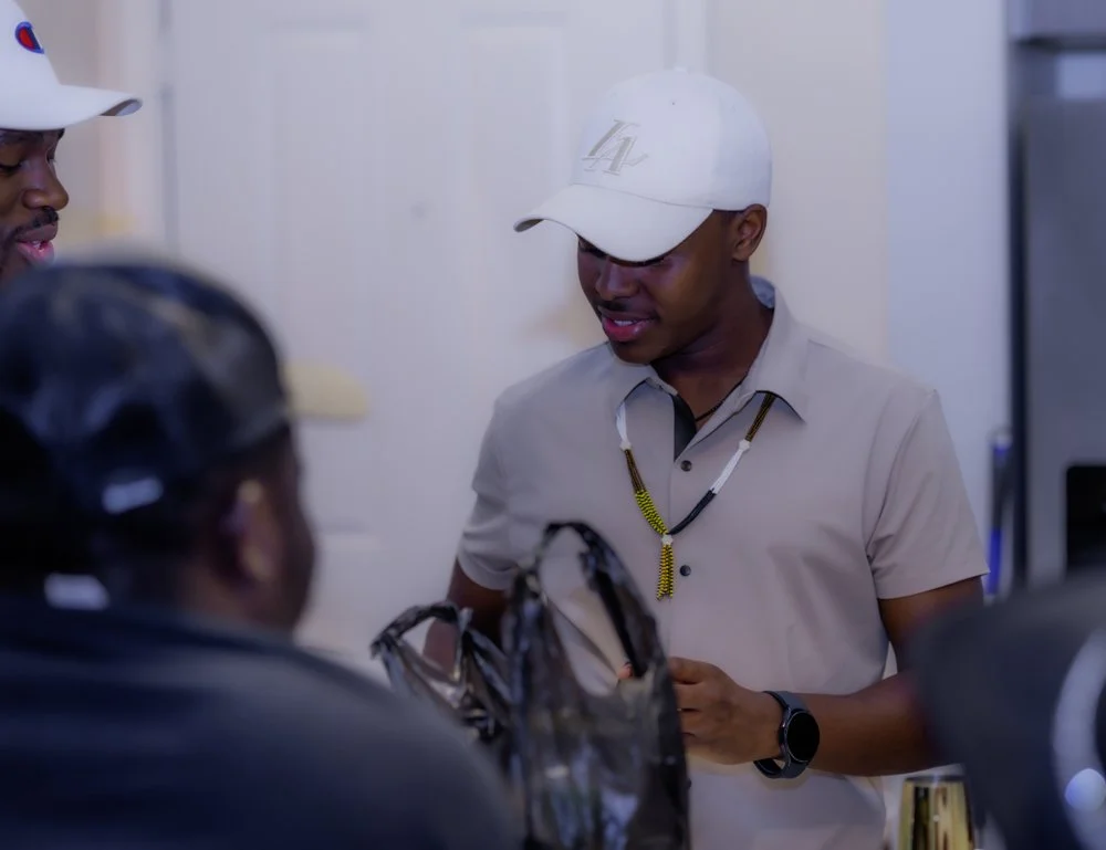 A group of people talking, including a man wearing a white baseball cap and beige shirt with a beaded necklace, in an indoor setting.