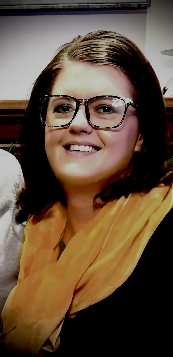 A woman smiling, wearing glasses, with short brown hair, and a yellow scarf around her neck.