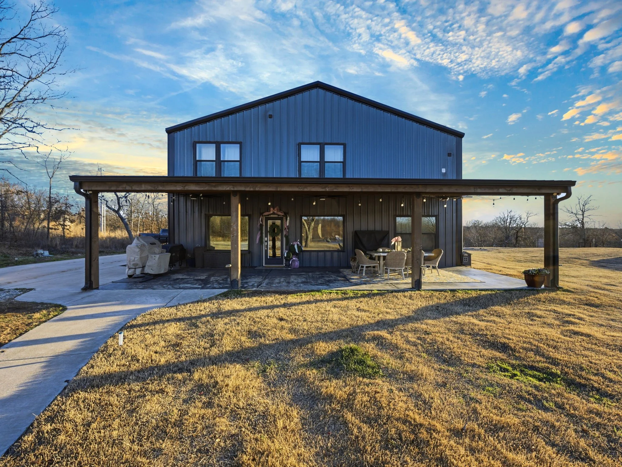 A two-story house with a metal blue exterior and a large front porch with seating. The house is surrounded by a lawn and driveway, with a few trees and a clear sky in the background.