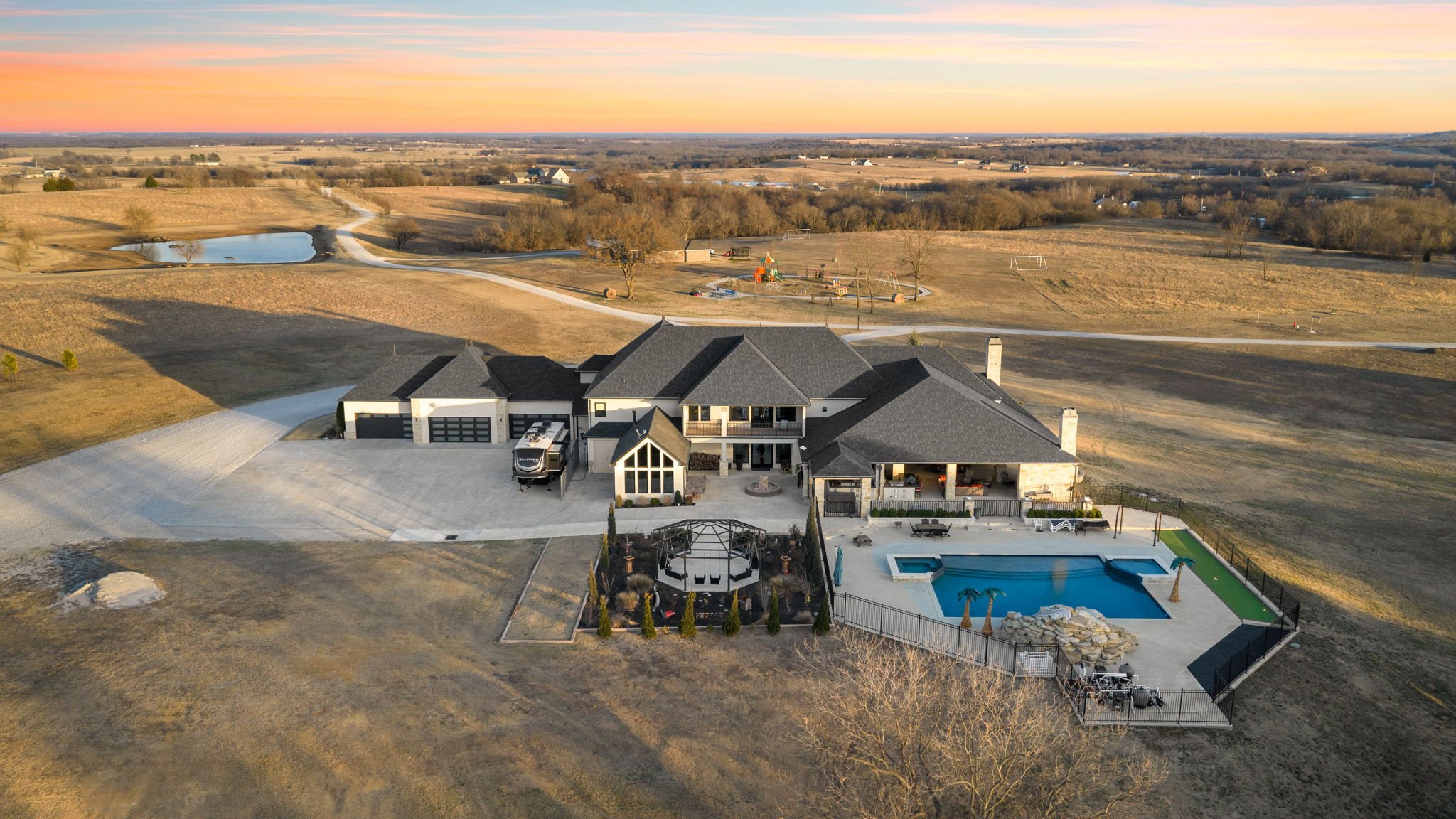 A large modern house with a pool, trampoline, and landscaped backyard, surrounded by open fields and a pond at sunset.