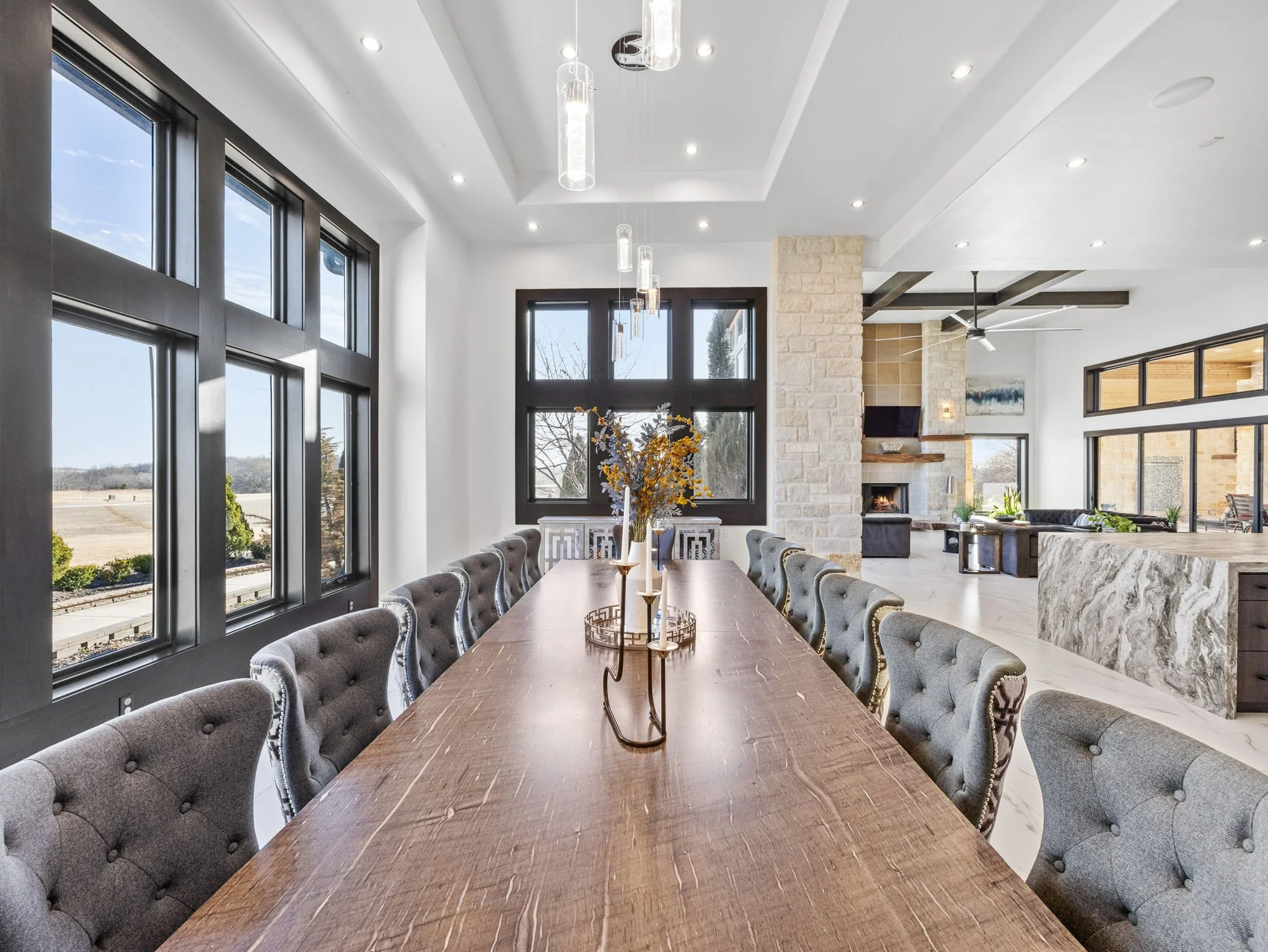 A spacious dining room with large windows, a long wooden dining table with 12 upholstered chairs, modern pendant lights, and a view of an outdoor landscape.