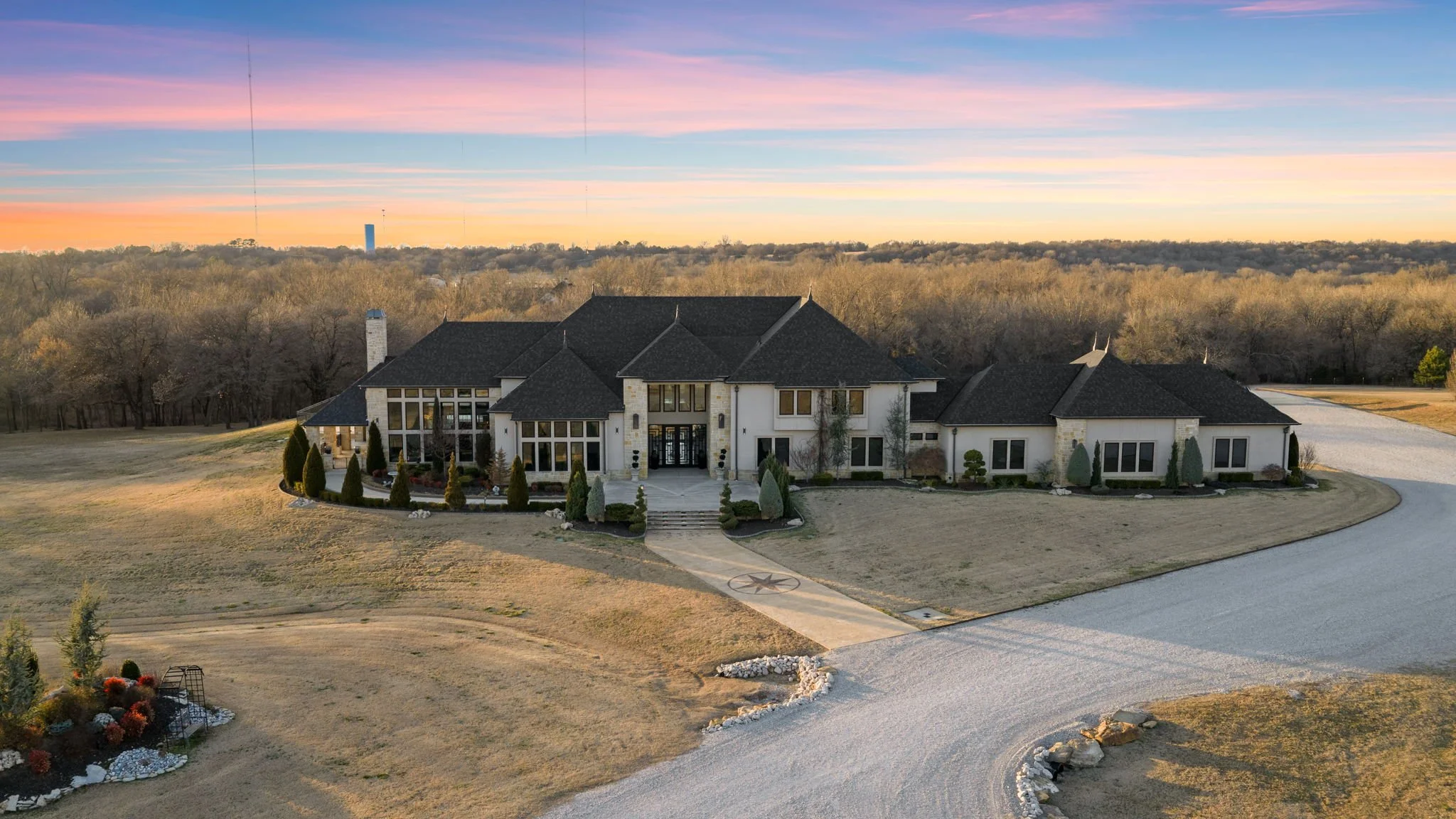 A large modern house with a dark roof and white walls, surrounded by a lawn with landscaped bushes and trees, set against a sunset sky.