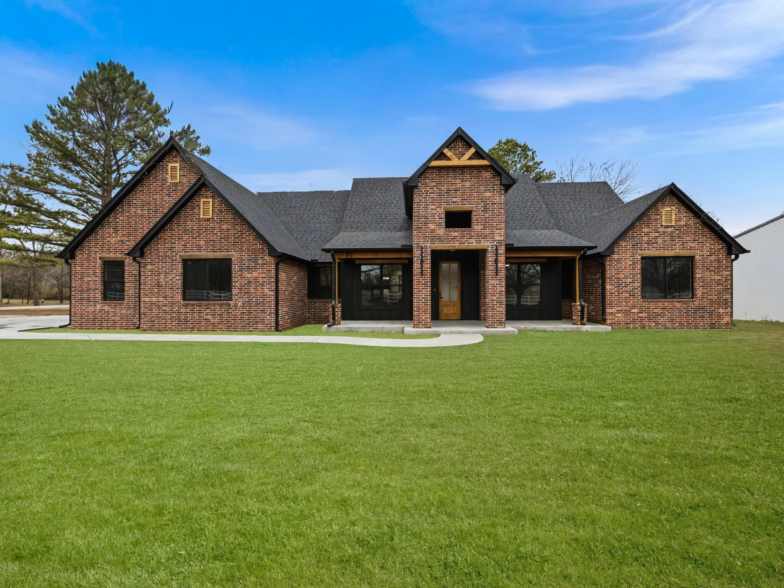 A large brick house with a dark roof, black window frames, and a wooden front door, set on a well-maintained green lawn under a blue sky.