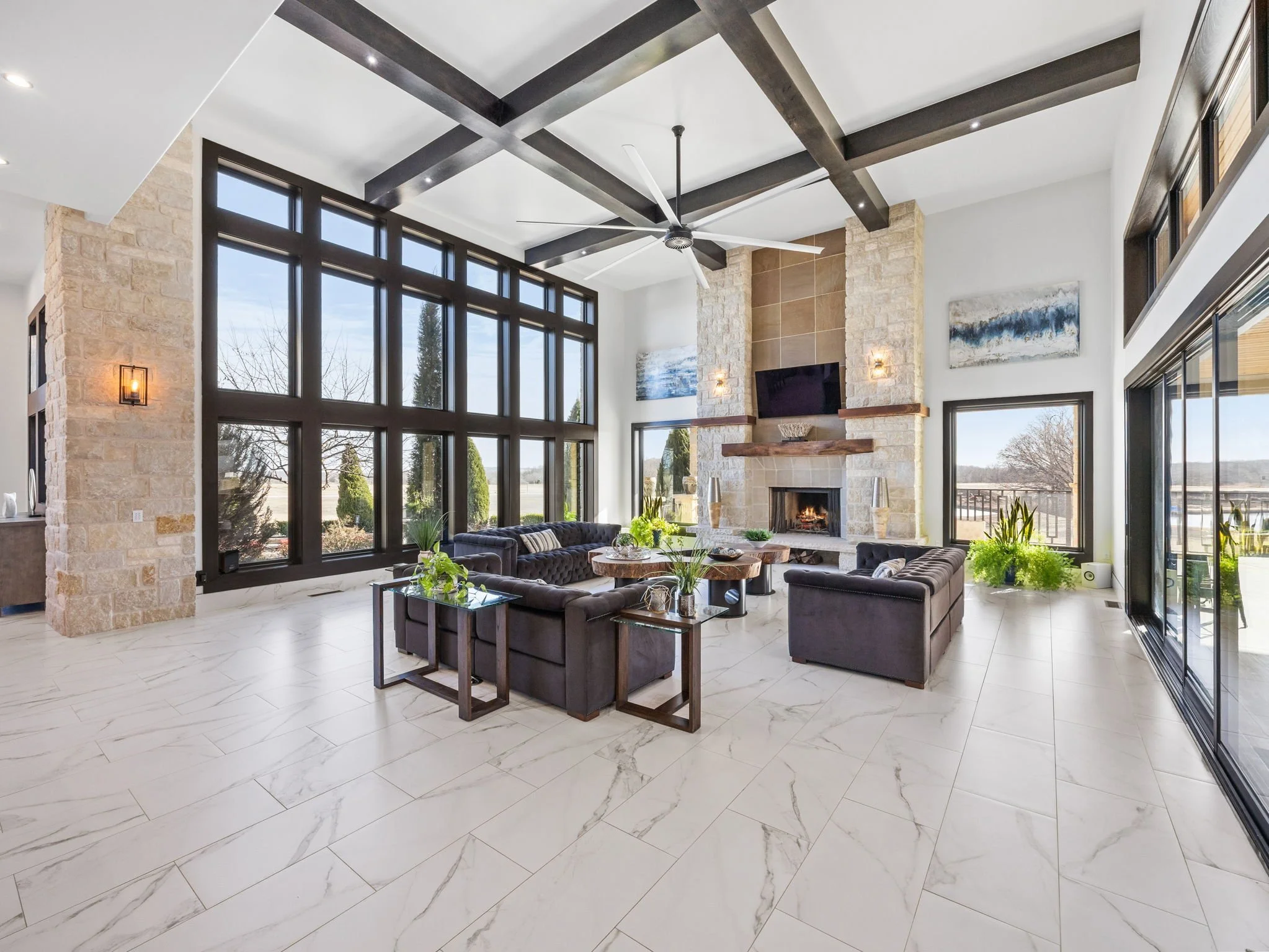 Spacious living room with large windows, a stone fireplace with a mounted TV, black sofas, coffee tables with plants, and artwork on white walls.